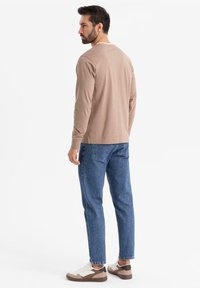 Camicia marrone chiaro a maniche lunghe con scollo tondo, abbinata a jeans blu e scarpe sportive con accenti marroni. Design minimalista, vestibilità casual.