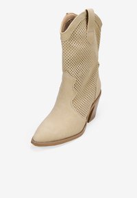 Botas de vaquero beige con parte superior perforada, punta afilada y diseño lateral curvado. Cuenta con un tacón bajo apilado y una textura de ante suave.