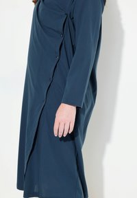 Abito blu scuro realizzato in tessuto morbido, con una chiusura frontale a bottoni, maniche lunghe e un design drappeggiato che crea texture e movimento.