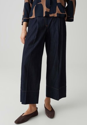 Personne portant un pantalon en jean large et foncé, associé à des chaussures plates marron et un haut noir et marron à motifs.