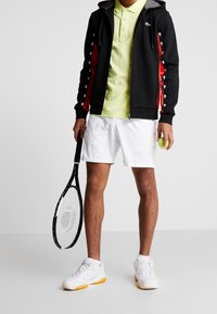 Svart zip-up hoodie med röda detaljer, ljusgrön polo, vita shorts och tennisskor. Håller en tennisracket och en tennisboll.