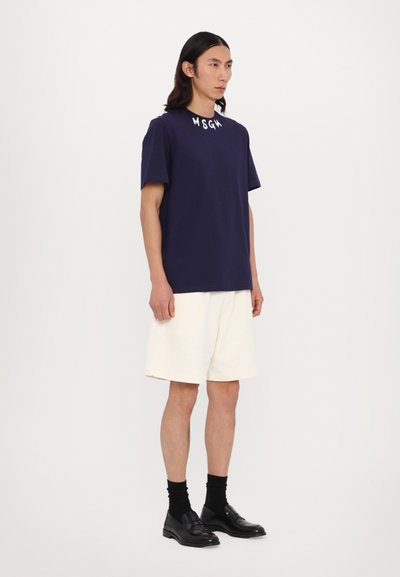 Tume t-särk, mille kaeluses on "MSGM" logo, koos kreemjatest lühikestest pükstest. Mudel kannab musti loafereid ja sokke, seistes valge taustaga.