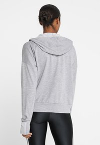 Vrouw met donker haar in een knot, gekleed in een lichtgrijze hoodie en zwarte leggings, van achteren gezien tegen een witte achtergrond.