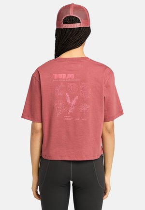 Kobieta z zaplecionymi włosami, ubrana w różowy t-shirt Timberland i siateczkową czapkę, czarne legginsy, zwrócona tyłem, ukazująca kwiatowe wzory i tekst na koszulce.