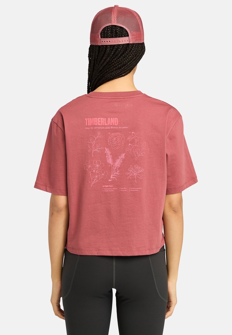 Femme aux cheveux tressés portant un t-shirt Timberland rose et une casquette en mesh, leggings noirs, vue de dos dévoilant des motifs floraux et du texte sur le t-shirt.