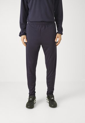 Pantalons de sport navy avec une taille élastique, coupe ajustée, texture lisse et jambes fuselées, associés à des chaussures de football noires avec des rayures blanches.