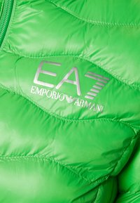 Zelená nadýchaná bunda s hladkou, polstrovanou texturou. Na hrudi se nachází stříbrné logo "EA7 Emporio Armani". Kompletní zipový design se švy.