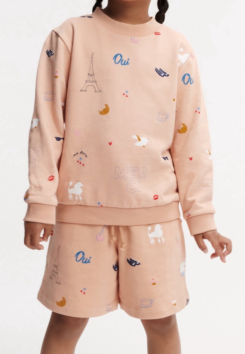 Enfant portant un sweat-shirt pêche et un short avec des impressions à thème français comprenant la tour Eiffel, des croissants, des oiseaux et le texte "Oui".