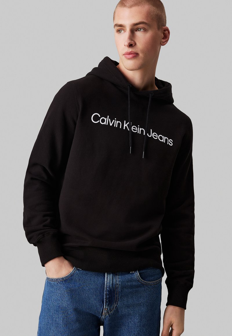 Sudadera negra con un gráfico frontal que dice "Calvin Klein Jeans", con capucha ajustable y puños de canalé, combinada con unos jeans azules.