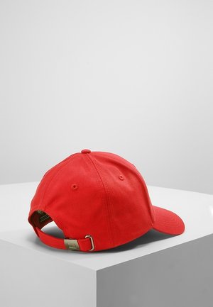 Czerwona czapka bejsbolowa z regulowanym paskiem i metalową sprzączką, spoczywająca na białym geometrycznym piedestale na jednolitym szarym tle.