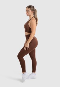 Een bruin sportbeha en bijpassende high-waisted legging gemaakt van rekbaar materiaal, met een strakke uitstraling. Witte sneakers maken de outfit compleet.
