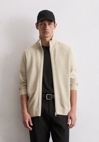 Beige ritsjack van gestructureerde stof met een hoge kraag, gecombineerd met een zwart T-shirt en een strakke zwarte broek, accessorized met een zwarte pet.