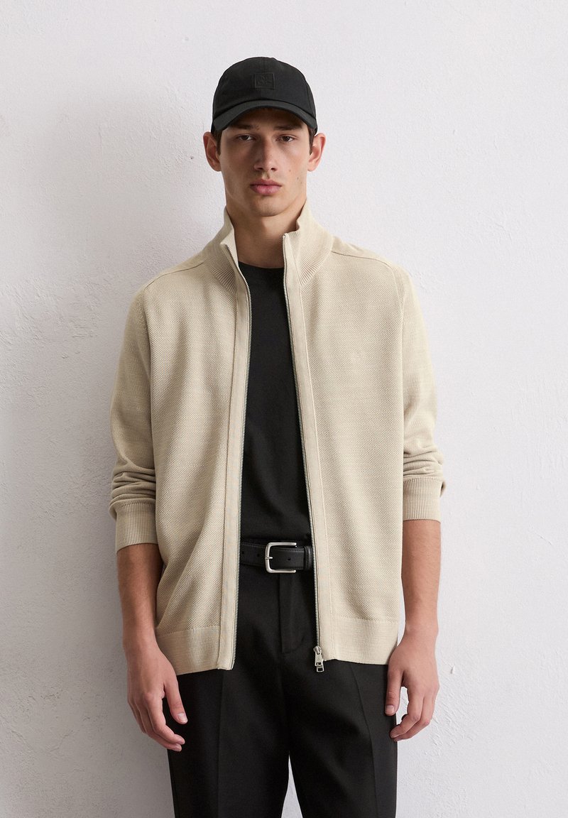 Beige ritsjack van gestructureerde stof met een hoge kraag, gecombineerd met een zwart T-shirt en een strakke zwarte broek, accessorized met een zwarte pet.