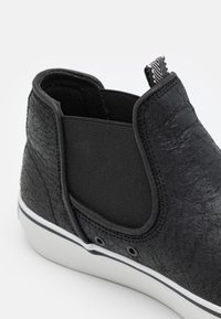 Botín negro de tobillo con cuero sintético texturizado y panel lateral elástico. Presenta una suela de goma blanca y una pequeña lengüeta con patrón a rayas.