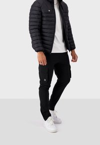 Giacca a vento nera con design trapuntato, abbinata a pantaloni cargo neri con tasche a pattina e sneaker bianche con dettagli perforati.