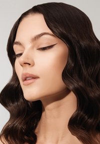 Primo piano di un volto con pelle liscia e chiara. Capelli scuri e ondulati incorniciano il viso. Le caratteristiche includono una linea di eyeliner affilata e labbra morbide.