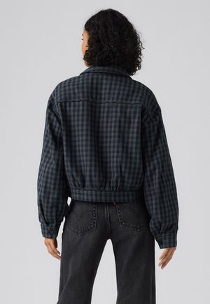 Personne aux cheveux bouclés de dos portant une veste à carreaux bleu foncé et noir et un jean noir sur un fond gris uni.