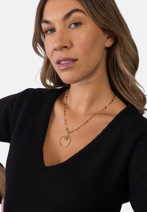 Woman with light brown hair wearing a black V-neck top and a gold necklace with a circular pendant featuring a small flower design.