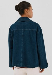 Czarna dżinsowa kurtka o luźnym kroju, z wyraźnym kołnierzem i widocznymi szwami. Posiada tylny karczek oraz długie rękawy z guzikami przy mankietach.