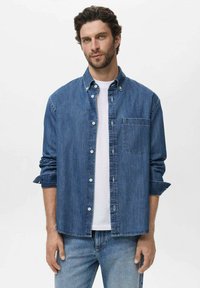 Camicia di denim con maniche lunghe, colletto a bottoni, taschino frontale e colore azzurro chiaro. Indossata sopra una t-shirt bianca a girocollo.