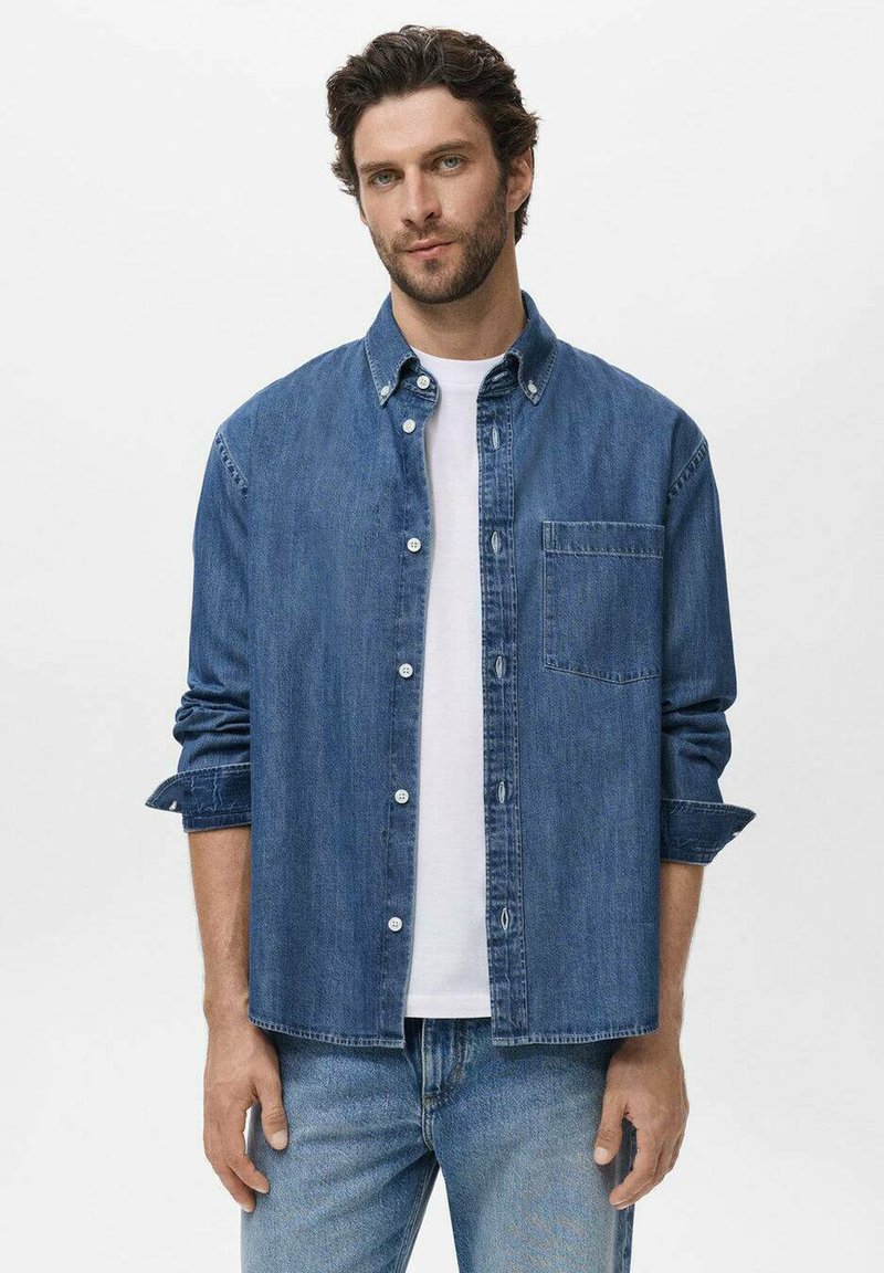Camicia di denim con maniche lunghe, colletto a bottoni, taschino frontale e colore azzurro chiaro. Indossata sopra una t-shirt bianca a girocollo.
