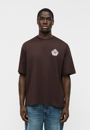 Mladý muž v tmavohnedom tričku s malým bielym ozdobným logom na ľavej strane hrude, v modrých džínsoch, stojaci na jednoduchom pozadí.