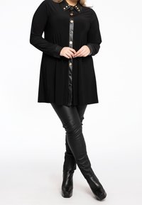 Schwarzes langärmliges Kleid mit spitzem Kragen und Metallknopf-Akzenten. Kombiniert mit eng anliegenden schwarzen Kunstlederhosen und klobigen schwarzen Stiefeln mit Absatz.