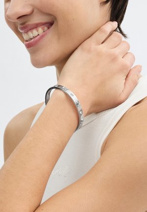 Zilveren metalen armband met een glad ontwerp, versierd met schedelaccenten en ingelegde strasssteentjes. Draagt op een pols tegen een neutrale achtergrond.