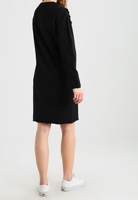 Svart, långärmad klänning med rak skärning och rund hals, med subtila detaljer vid axlarna. Bärs med vita sneakers.