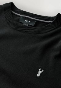 Juodas megztinis su raukšlėta iškirpte. Kairėje pusėje yra mažas pilkas išsiuvinėtas elnio logotipas. Medžiaga atrodo minkšta ir tekstūruota.