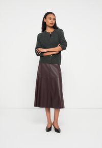 Donna in piedi con le braccia incrociate, che indossa un maglione grigio scuro con bottoni, una gonna di pelle marrone scuro a metà lunghezza e scarpe nere a punta.