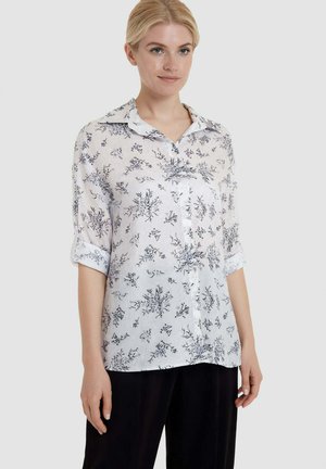 Donna che indossa una camicia bianca a maniche lunghe con motivi floreali neri e pantaloni neri, in piedi davanti a uno sfondo chiaro e semplice.
