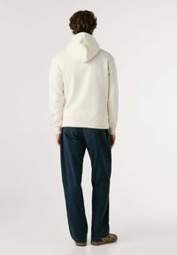 Man die met zijn rug naar de camera staat, gekleed in een off-white hoodie, donkerblauwe broek en bruine schoenen, tegen een eenvoudige lichte achtergrond.
