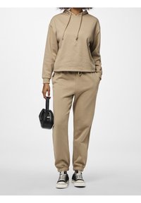 Felpa con cappuccio beige e pantaloni jogger coordinati con polsini elasticizzati, abbinati a sneakers nere e una piccola borsa nera. Tessuto morbido, vestibilità casual.