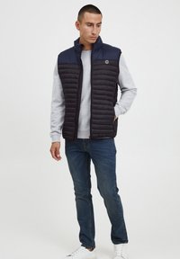 Navy Puffer-Weste mit gestepptem Design, hohem Kragen und Reißverschluss. Getragen über einem hellgrauen Sweatshirt, kombiniert mit blauen Jeans und weißen Sneakers.