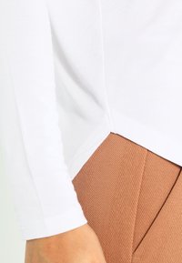 Camicia bianca a maniche lunghe con una trama a coste, dotata di orlo con spacco laterale. Abbinata a pantaloni marroni texturizzati a vita alta.