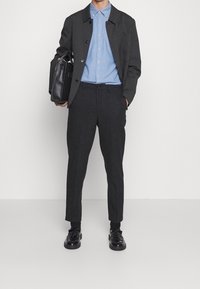 Svart kappa över en ljusblå skjorta, tillsammans med mörka byxor och svarta skor. Tillbehören inkluderar en svart väska. Släta texturer och strukturerad design.