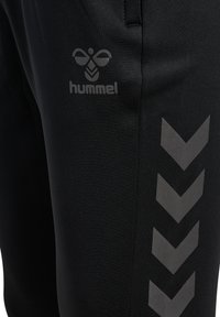 Schwarze Sporthosen mit glatter Textur, versehen mit einem grauen Hummel-Logo und einem Chevron-Muster an der Seite in einem dunkleren Grauton.