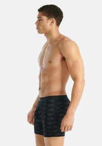 Svalnatý muž s krátkými kudrnatými vlasy, oblečený v černých boxerách se vzorem, stojící v profilu na neutrálním světlém pozadí.