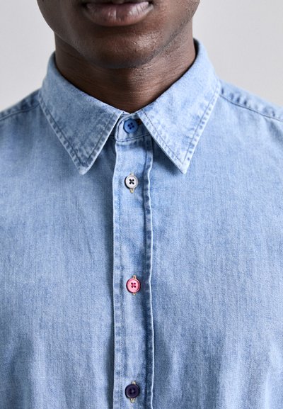 Camisa de mezclilla azul claro con cuello puntiagudo, que presenta botones coloridos: azul, rosa, blanco y morado oscuro a lo largo de la tapeta.
