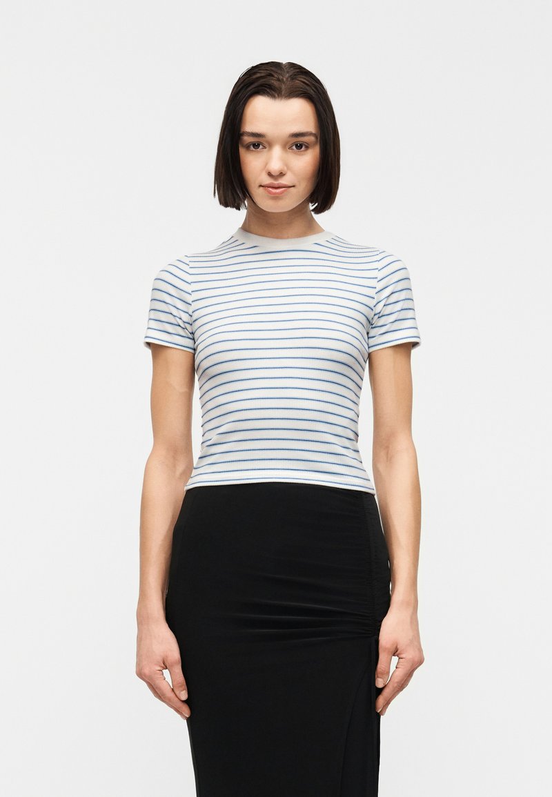 Femme jeune aux cheveux bruns raides portant un haut blanc à manches courtes avec de fines rayures horizontales bleues et une jupe noire.