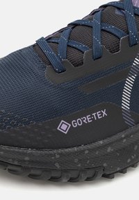Zapatilla de trail running con parte superior de malla en un oscuro azul marino, acentos sintéticos en negro, detalles en púrpura y una suela de goma texturizada que muestra el logo de GORE-TEX.