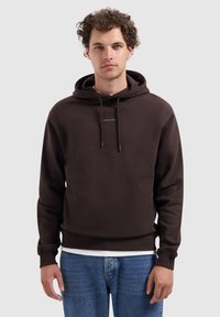 Man met krullend haar, gekleed in een donkerbruine hoodie met de tekst "PURE PATH", lichtblauwe spijkerbroek en een wit shirt eronder, staand voor een witte achtergrond.