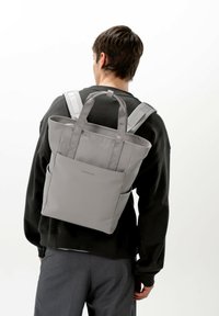 Grå axelväska med dubbla handtag och vadderade remmar. Texturerad tyg, rektangulär form med externa sidofickor för förvaring.