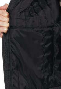 Schwarze isolierte Jacke mit glattem, glänzendem Innenfutter, gesteppten Nähten und einer seitlichen Tasche. Eine Hand greift den Stoff.