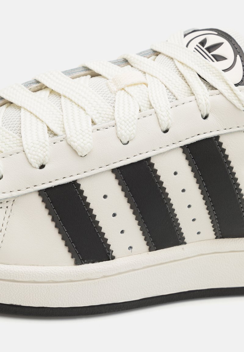 Baskets blanches avec des rayures en cuir noir et des perforations, semelle en caoutchouc texturé et logo de marque bien visible sur la languette. Surface lisse.