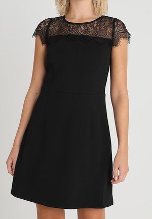 Kobieta ubrana w czarną sukienkę o kroju A z koronkowymi, krótkimi rękawkami oraz koronkowym karczkiem na górnej części klatki piersiowej i na ramionach.