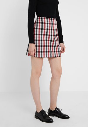 Frau trägt ein schwarzes Langarmoberteil, einen rot-schwarz-weiß karierten Rock und schwarze Leder-Schnürschuhe, steht vor einem weißen Hintergrund.