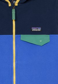 Veste en polaire bleue avec des blocs de couleurs bleu marine, bleu clair et vert. Dotée d'une fermeture éclair jaune et d'une poche à rabat avec bouton-pression. Logo Patagonia affiché.