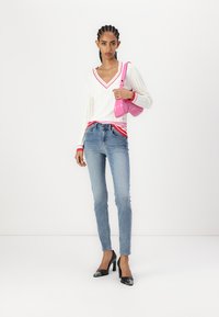 Maglione bianco a coste con scollo a V rosso e polsini rossi, abbinato a jeans in denim blu e scarpe con tacco nere. Borsa rosa come accessorio a contrasto.